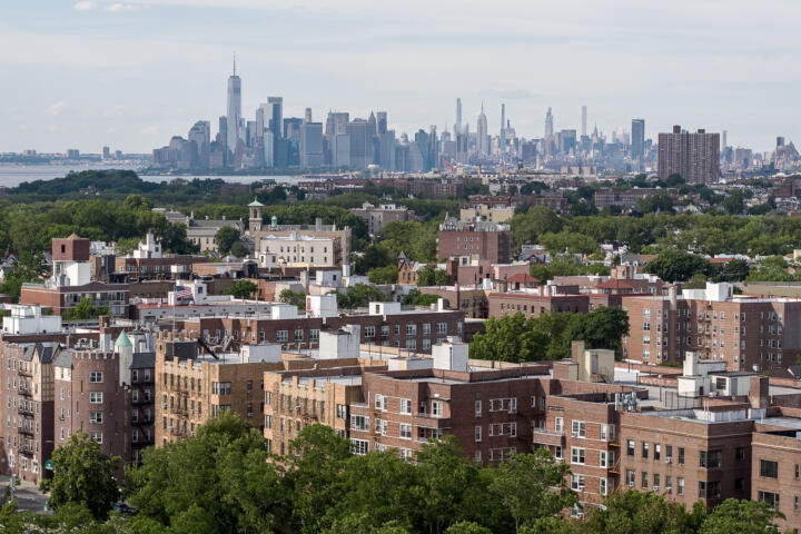 A wide view of a Brooklyn neighborhood with rows of mid-rise brick apartment buildings in the foreground, interspersed with green trees. In the background, the Manhattan skyline rises across the East River, with iconic skyscrapers like One World Trade Center visible under a partly cloudy sky.
