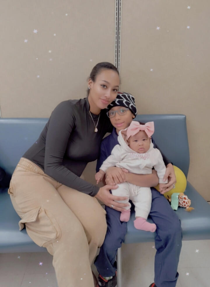 A person sits on a blue bench with two children in their lap. One child wears glasses and a patterned knit hat, while the baby wears a light-colored outfit with a large pink bow headband. The group is indoors against a neutral-colored wall with star-like filters added to the photo.