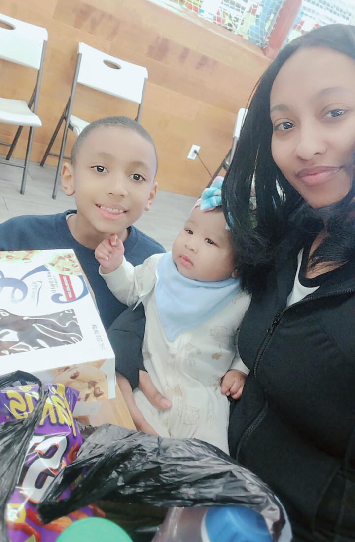 A smiling adult sits with two children at a table. The older child, wearing a navy blue sweatshirt, smiles at the camera while the baby, dressed in a white onesie and a blue bow headband, rests in the adult's arms. The table in front of them holds packaged cookies, snacks, and a drink bottle. The background shows white folding chairs and a wooden wall.