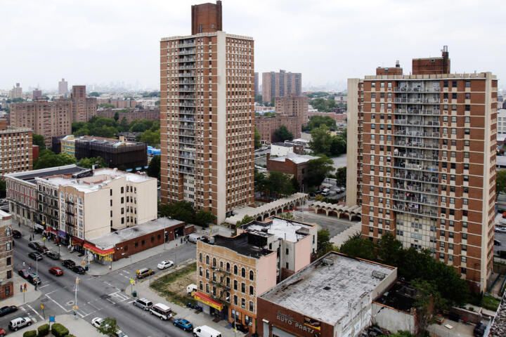 Aerial view of Brownsville in Brooklyn, New York, showing several tall public housing towers surrounded by mid-rise apartment buildings and low-rise storefronts along busy streets. The neighborhood appears densely built with patches of trees and green space. The sky is overcast, and the distant New York City skyline is faintly visible in the background.