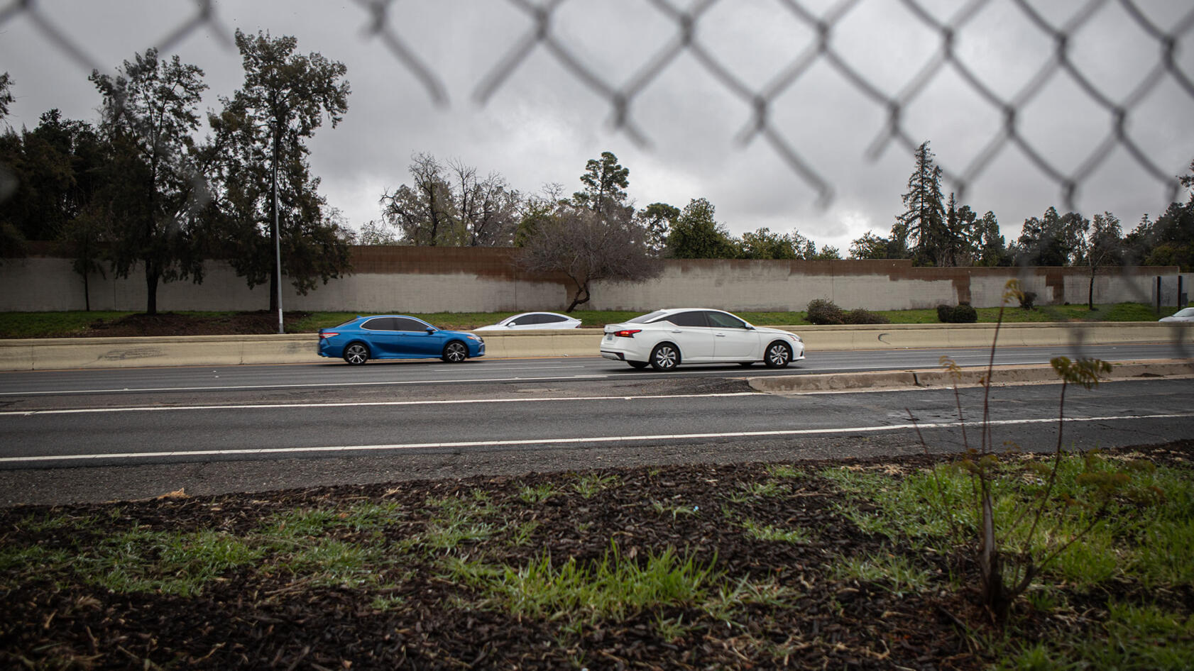 A side-view, as seen through a partially broken wire fence, of cars zooming through a highway on a cloudy day.