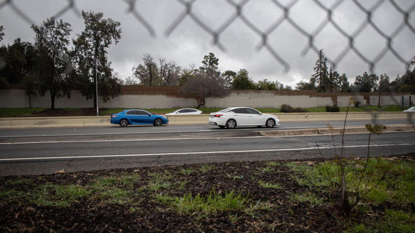 A side-view, as seen through a partially broken wire fence, of cars zooming through a highway on a cloudy day.