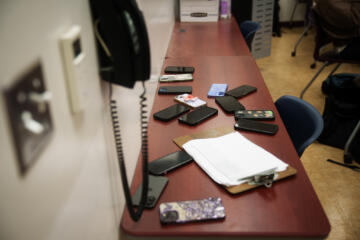 A group of smartphones is spread across a wooden desk in a classroom or office setting. A clipboard with a sign-in sheet rests among the phones. Nearby chairs and part of a person sitting at a desk are visible in the background. A wall-mounted phone and other typical office fixtures are seen along the left edge of the frame.