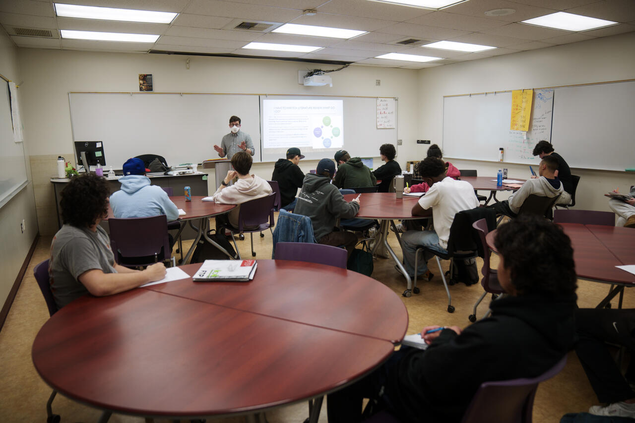 A classroom full of students sits at round tables while an instructor at the front of the room gives a presentation. The instructor wears a face mask and gestures with their hands as a slide projected behind them explains how to write a literature review. Most students are engaged, taking notes or listening attentively. The classroom is brightly lit with overhead lights and whiteboards lining the walls.