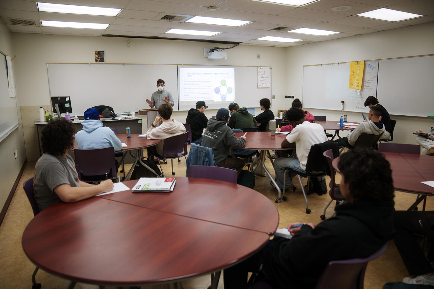 A classroom full of students sits at round tables while an instructor at the front of the room gives a presentation. The instructor wears a face mask and gestures with their hands as a slide projected behind them explains how to write a literature review. Most students are engaged, taking notes or listening attentively. The classroom is brightly lit with overhead lights and whiteboards lining the walls.