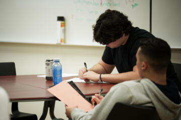 Two students sit at a round classroom table focused on writing. One is writing on white paper while the other holds a bright orange sheet with notes. A bottle of water and an energy drink sit on the table. A whiteboard with faint writing and a canister of cleaning spray are visible in the background.