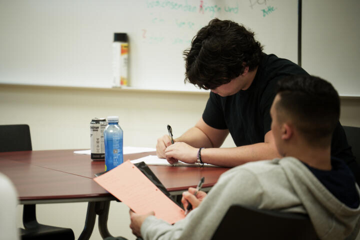 Two students sit at a round classroom table focused on writing. One is writing on white paper while the other holds a bright orange sheet with notes. A bottle of water and an energy drink sit on the table. A whiteboard with faint writing and a canister of cleaning spray are visible in the background.