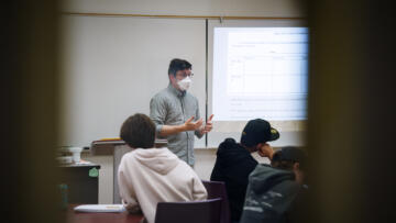 An instructor wearing a mask stands at the front of a classroom, gesturing while speaking to students seated at round tables. A projected slide behind the instructor displays a chart titled “Creating Your Synthesis Chart,” outlining how to organize sources and main ideas. The image is framed by two blurred vertical objects in the foreground, giving the perspective of someone observing through a narrow space.