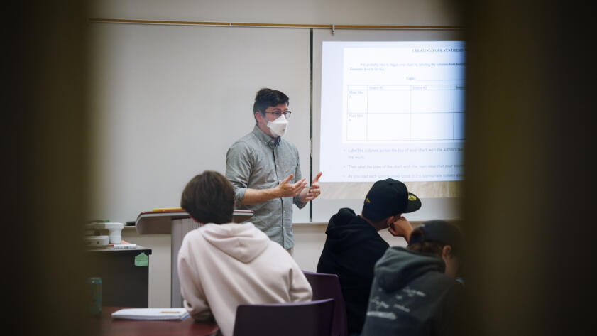 An instructor wearing a mask stands at the front of a classroom, gesturing while speaking to students seated at round tables. A projected slide behind the instructor displays a chart titled “Creating Your Synthesis Chart,” outlining how to organize sources and main ideas. The image is framed by two blurred vertical objects in the foreground, giving the perspective of someone observing through a narrow space.