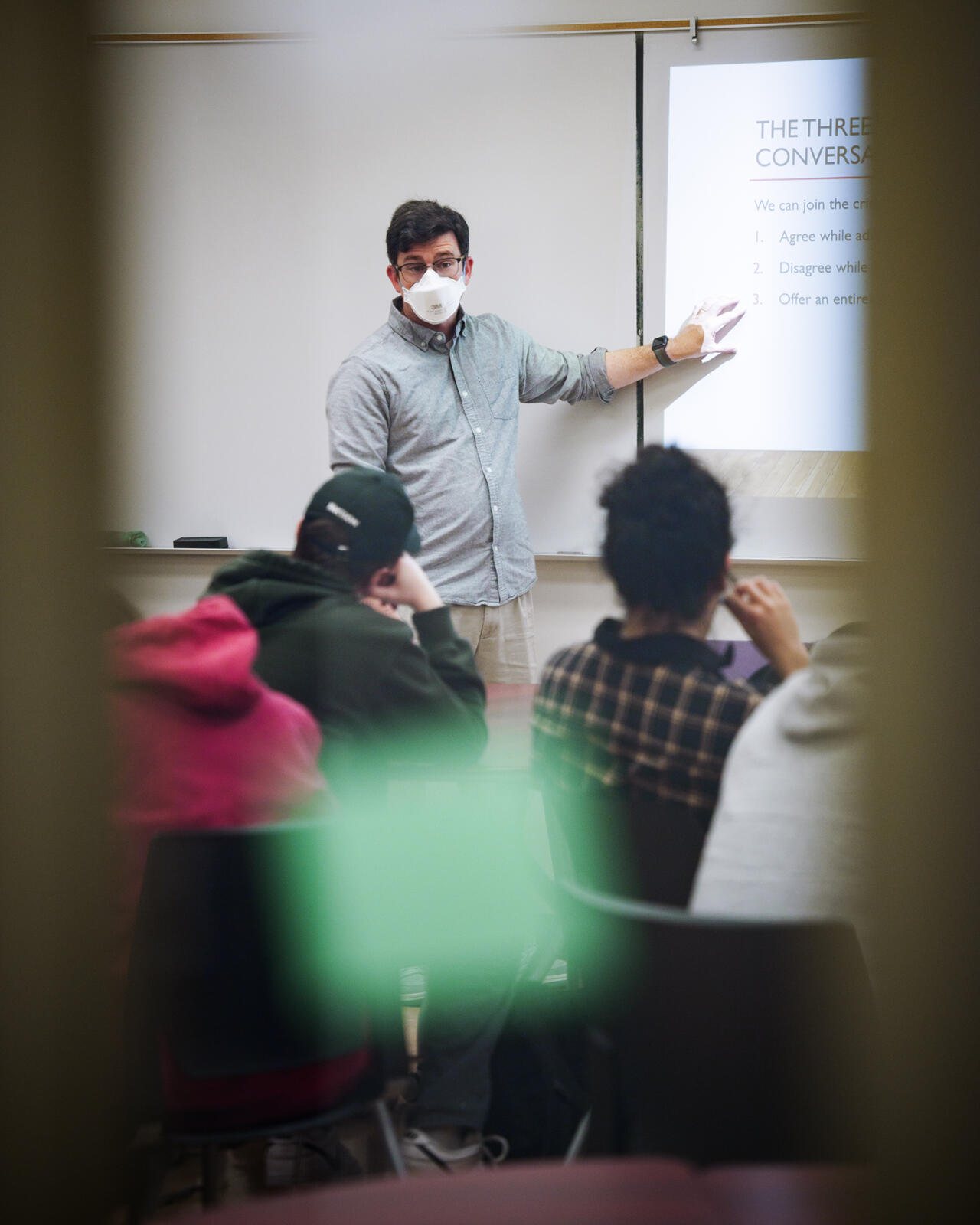 A masked instructor stands at the front of a classroom, pointing to a projected slide titled "The Three Conversations." The instructor appears to be mid-lecture, facing a group of seated students. The image is framed through a window or door with blurred green and yellow shapes in the foreground, adding a sense of candid observation.
