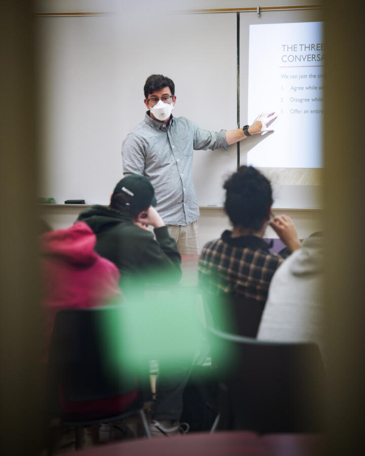 A masked instructor stands at the front of a classroom, pointing to a projected slide titled "The Three Conversations." The instructor appears to be mid-lecture, facing a group of seated students. The image is framed through a window or door with blurred green and yellow shapes in the foreground, adding a sense of candid observation.