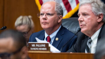 A person, with short white hair and wearing a blue suit, sits in front of a dais alongside other lawmakers during a hearing. A portion of an American flag can be seen in the background.
