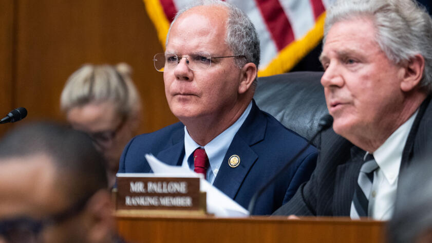 A person, with short white hair and wearing a blue suit, sits in front of a dais alongside other lawmakers during a hearing. A portion of an American flag can be seen in the background.