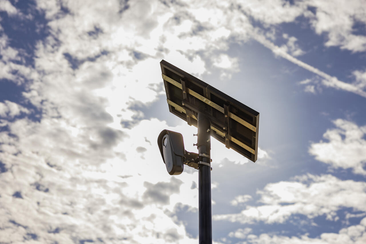 A surveillance camera mounted on a tall pole with a solar panel above it, set against a partly cloudy sky with the sun glowing behind the clouds.
