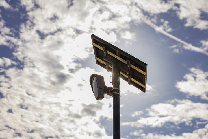 A surveillance camera mounted on a tall pole with a solar panel above it, set against a partly cloudy sky with the sun glowing behind the clouds.