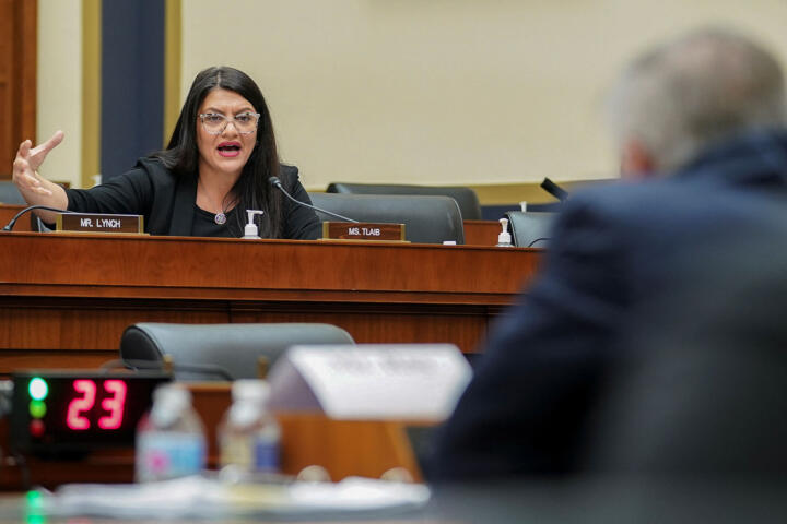 A lawmaker, with long black hair and wearing reading glasses, sits in front of a dais while speaking into a microphone as they ask questions to a person sitting in front of them during a congressional hearing in a legislative chamber.