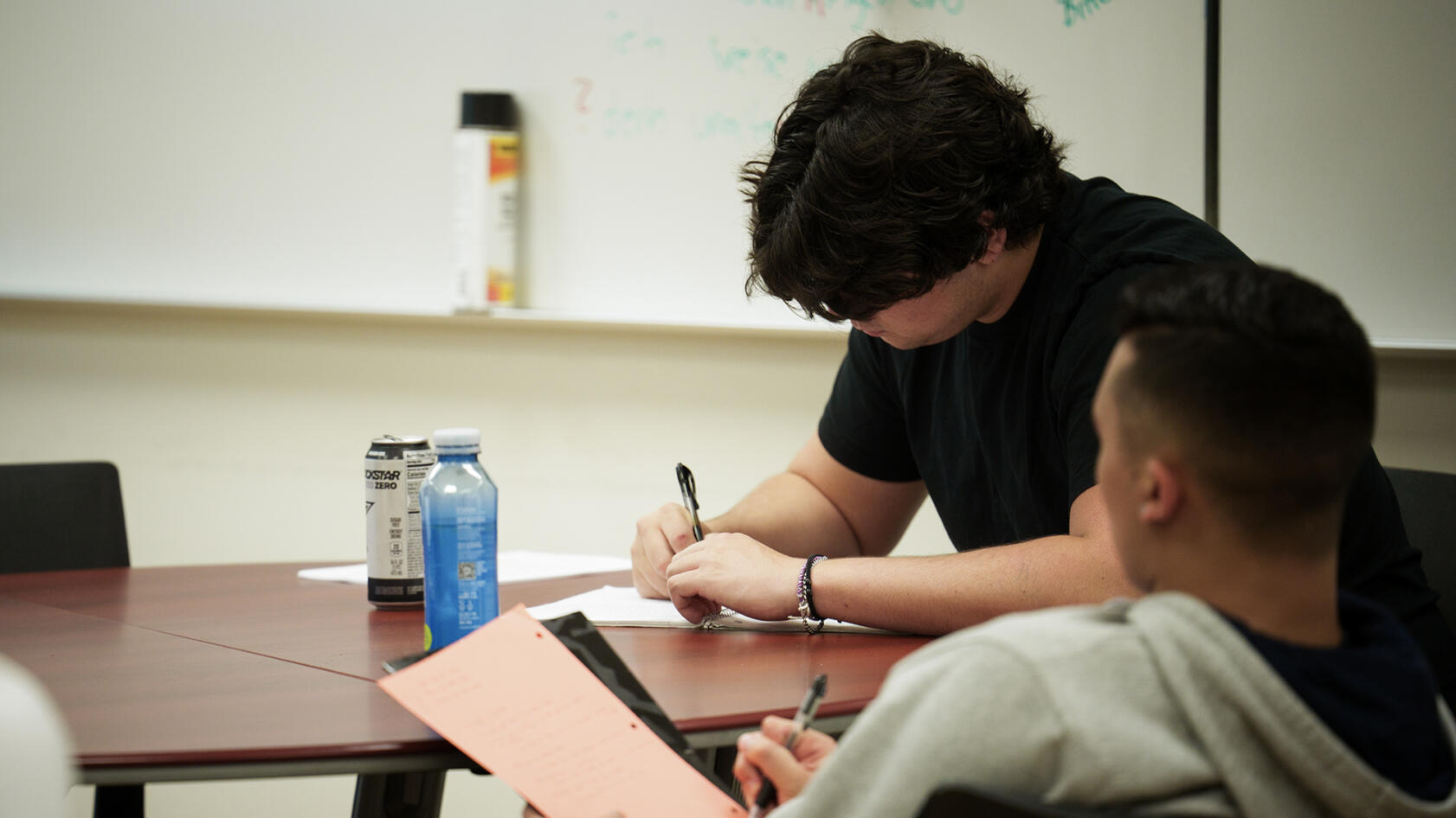 Two individuals sit at a table in a classroom, writing on paper with pens. One person with dark, wavy hair and a black shirt focuses on their work, while the other, wearing a gray hoodie, holds a notebook with a bright orange sheet. A can of energy drink and a blue bottle sit on the table. A whiteboard with faint writing is visible in the background.