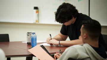 Two individuals sit at a table in a classroom, writing on paper with pens. One person with dark, wavy hair and a black shirt focuses on their work, while the other, wearing a gray hoodie, holds a notebook with a bright orange sheet. A can of energy drink and a blue bottle sit on the table. A whiteboard with faint writing is visible in the background.