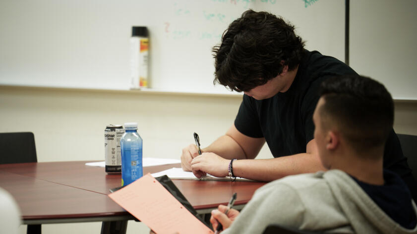 Two individuals sit at a table in a classroom, writing on paper with pens. One person with dark, wavy hair and a black shirt focuses on their work, while the other, wearing a gray hoodie, holds a notebook with a bright orange sheet. A can of energy drink and a blue bottle sit on the table. A whiteboard with faint writing is visible in the background.