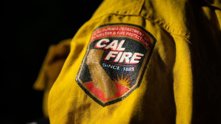 A close-up of a yellow Cal Fire uniform with a detailed embroidered patch on the shoulder. The patch features the shape of California, a stylized flame, and text reading “CAL FIRE” and “Since 1885.” The words “California Department of Forestry & Fire Protection” are visible along the top of the patch. The fabric is lit with strong contrast, emphasizing its texture and stitching.