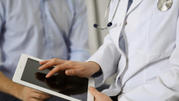 A person in a white medical coat with a stethoscope uses a tablet, while another individual in a light blue shirt sits nearby. The interaction suggests a consultation or discussion, with the focus on the hands and device rather than faces.