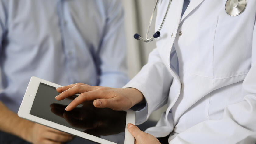 A person in a white medical coat with a stethoscope uses a tablet, while another individual in a light blue shirt sits nearby. The interaction suggests a consultation or discussion, with the focus on the hands and device rather than faces.