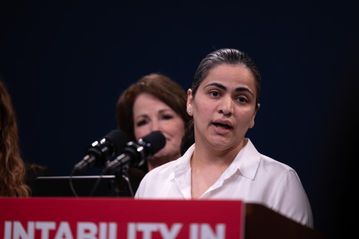 State Senator Aisha Wahab speaks at a podium with other lawmakers in the distance behind her.