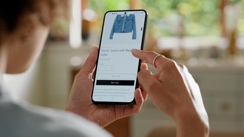 A person uses a smartphone to browse an online clothing store. The screen displays a product page for a denim jacket with a shearling collar priced at $219. The individual is about to tap the "Buy it now" button, with one hand holding the phone and the other poised to make the selection. The background is softly blurred, suggesting a home setting with natural light filtering in through windows.