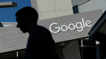 A tilted view of a person's silhouette as they walk in front ofa gray building with the Google logo in front, on a clear day.