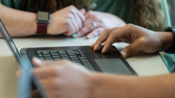A close-up view of a student's hands on a laptop keyboard. A teacher's hands can be seen nearby resting on the student's desk.