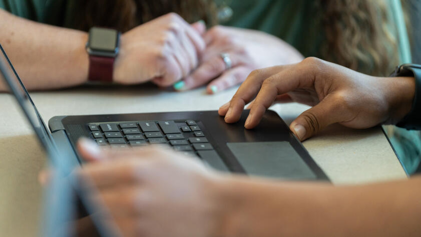 A close-up view of a student's hands on a laptop keyboard. A teacher's hands can be seen nearby resting on the student's desk.