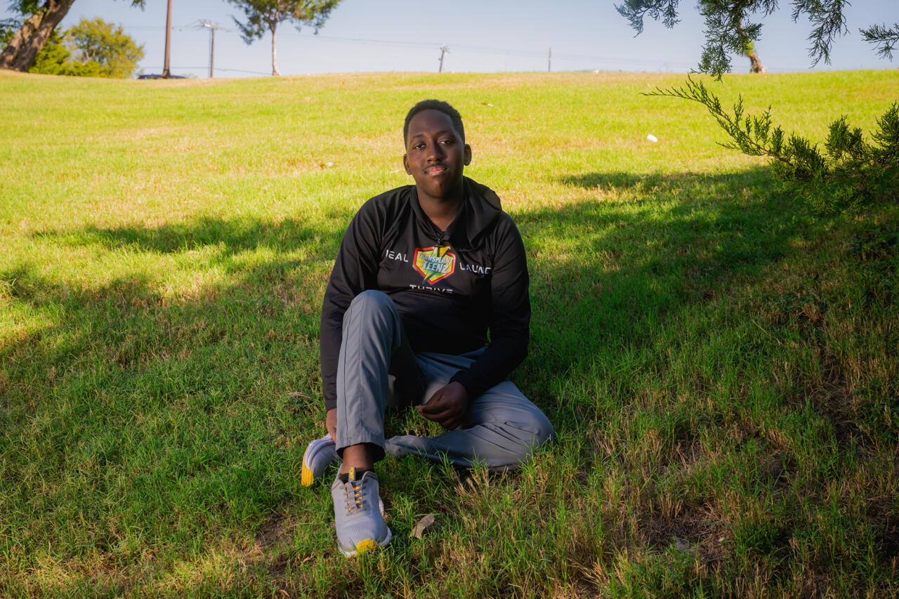 A person, wearing a black sweater and gray pants, sits on a grass hill shaded by a small tree on a clear day.