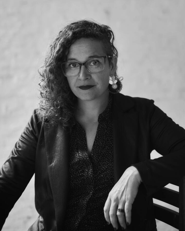 A person with curly hair on one side and a shaved undercut on the other sits angled toward the camera, wearing glasses, a patterned shirt, and a dark blazer. They have one arm resting on the back of a chair and look calmly at the viewer in this black-and-white portrait.