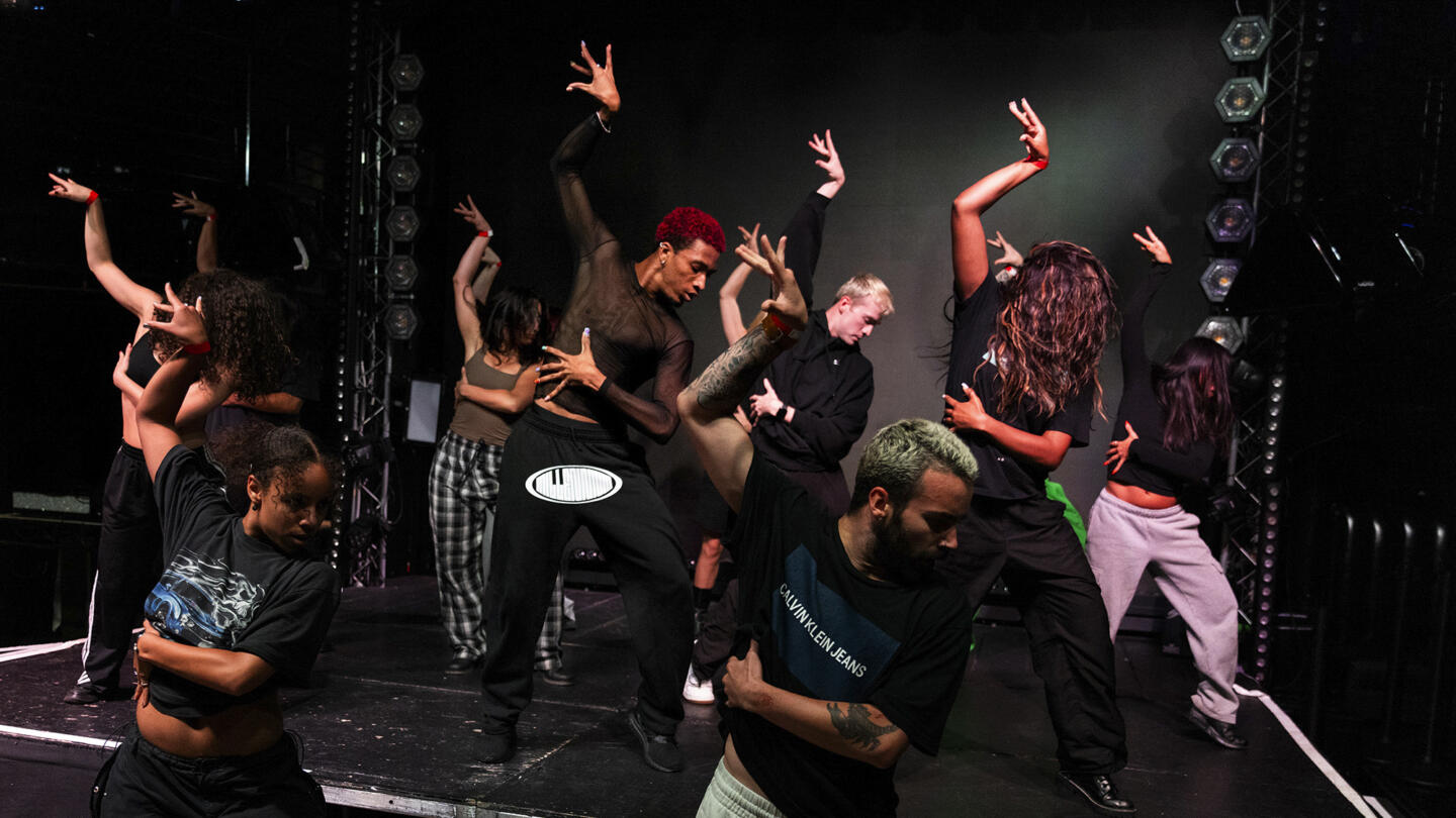 A dancing group poses in sync, their arms arched, standing in formation on a stage in a dark dance studio. In the background is a black backdrop with LED lights hanging on metal railing rigs.