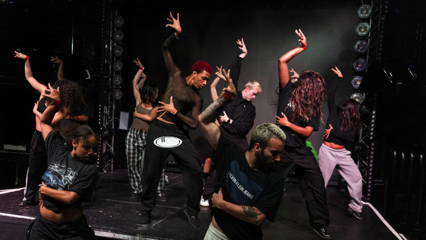 A dancing group poses in sync, their arms arched, standing in formation on a stage in a dark dance studio. In the background is a black backdrop with LED lights hanging on metal railing rigs.