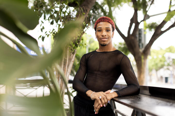 A person wearing a black mesh t-shirt leans against a wooden railing as they pose for a photo, with greenery in the background poking into the frame.