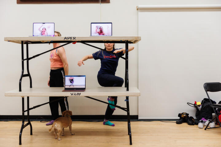 Two people are dancing behind two tables stacked on top of each other, with laptops on them, displaying them on the screens inside a dance studio. A dog can be seen walking beneath the table on hardwood floors, with a pile of bags and shoes next to a chair on the opposite side of the frame.
