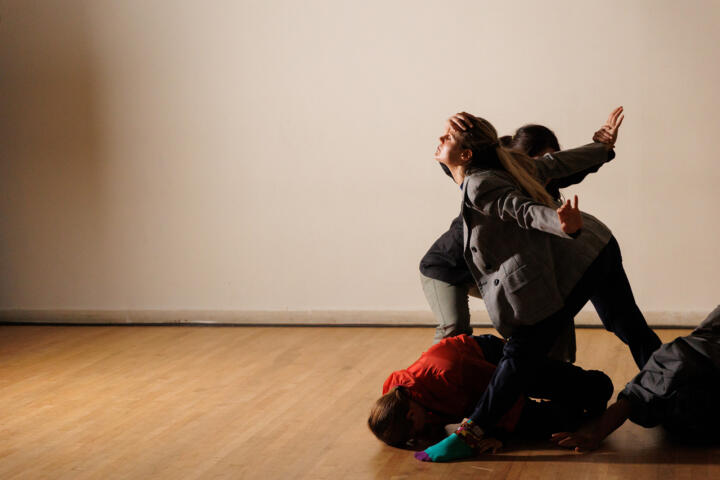 Three people arranged in a dance routine inside a plain-colored dance studio. One person has their back arched as they are in the middle of their dance routine, while another lies directly underneath them.