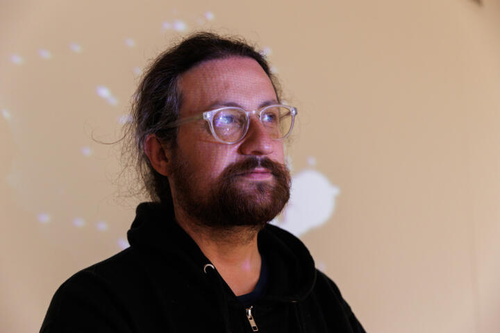 A person with glasses and a beard stands in a dance studio while a project shines on their face and reflects glare off their glasses' lenses.