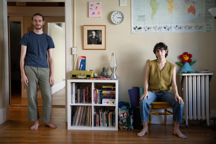 Two people pose for a photo near the wall and doorway of a room in an apartment. One is standing in the doorway while the other is sitting on a wooden chair. The room in the background is colorfully decorated with children's toys and a world map hung on the wall.