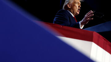 A tilted view of former U.S. President Donald Trump standing in front of a podium as he speaks from a stage decorated in blue, white and red colors.
