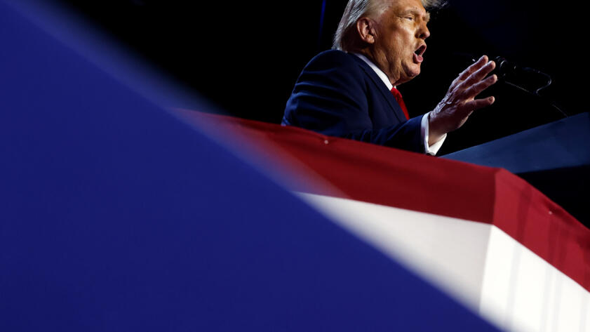 A tilted view of former U.S. President Donald Trump standing in front of a podium as he speaks from a stage decorated in blue, white and red colors.