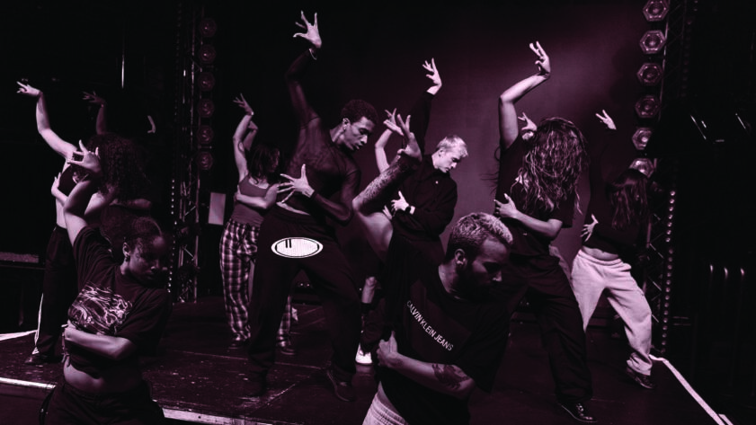Pink-tinted photograph of a dancing group posing in sync, their arms arched, standing in formation on a stage in a dark dance studio; the background is a black backdrop with LED lights hanging on metal railing rigs
