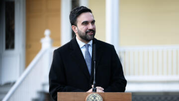A person in a dark coat and light blue tie speaks at a wooden podium outdoors, smiling slightly as a microphone stands in front of them, with the pale yellow facade and white railing of a government building blurred in the background.