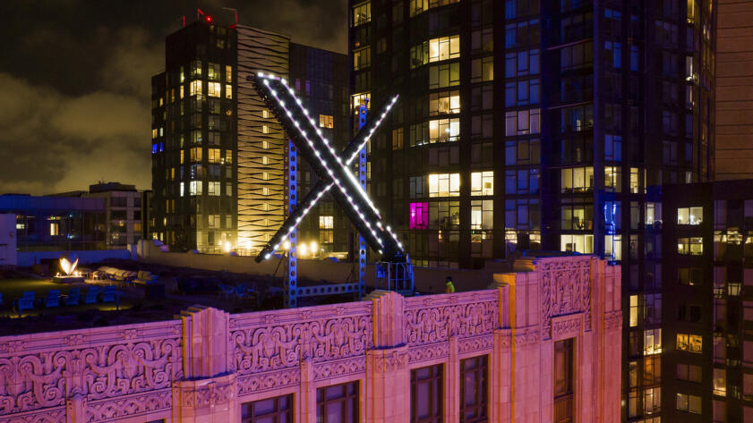 A view of workers on top of a building installing lighting on an "X" sign, with the windows of nearby buildings and the cityscape in the background.