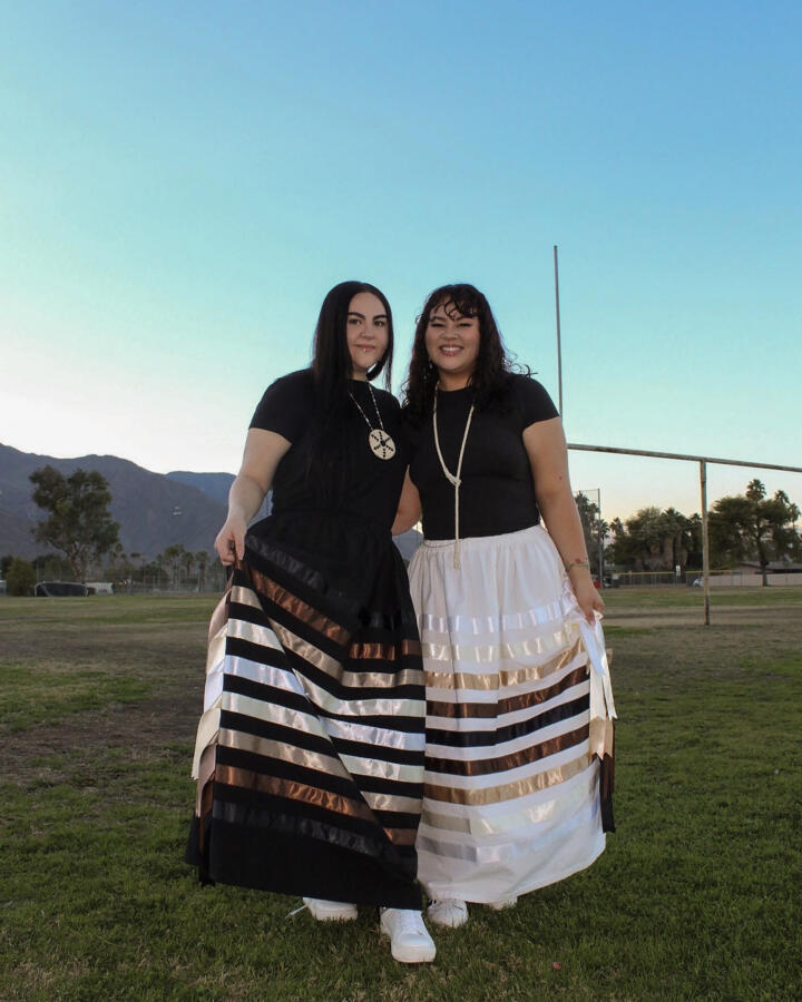 Two people stand side by side on a grassy field at dusk, wearing black tops and long skirts with horizontal bands of black, white, and metallic tones, smiling toward the camera with mountains and an open sky in the background.