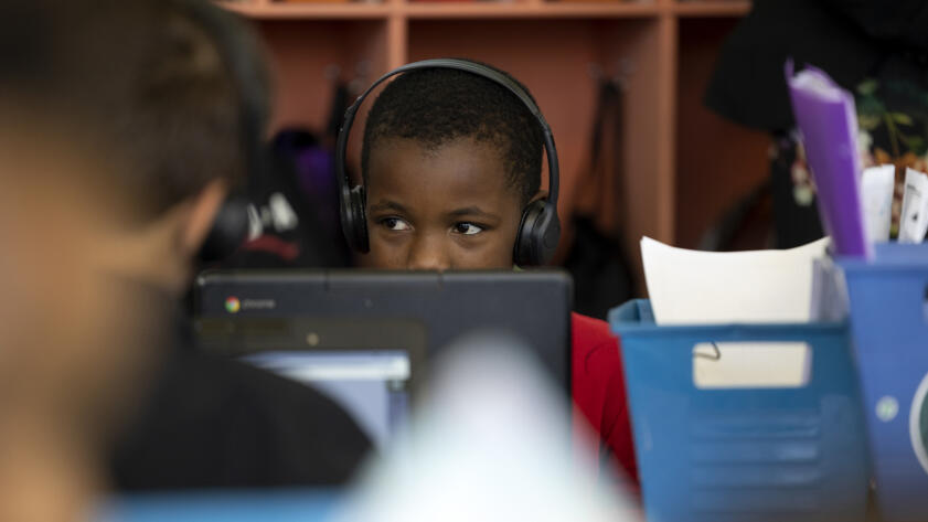 A young student sits at a table, headphones on, looking at a computer screen in a classroom. The students head peals over the top of the screen as their attention is focused on the computer.