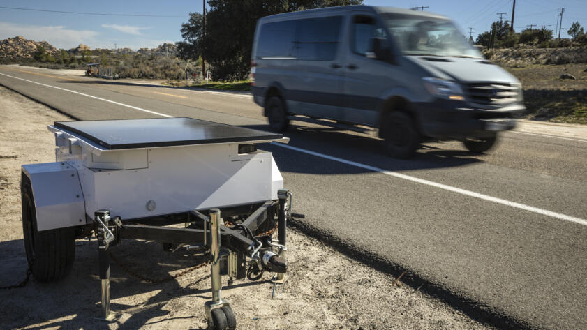 A roadside device mounted on a small trailer sits on the shoulder beside a two-lane road as a blurred van drives past, with utility poles, trees, and rocky hills in the background under a clear sky.