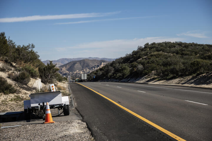 A portable roadside camera trailer sits on the shoulder beside a highway stretching through low, brush-covered hills, with a traffic cone placed near its hitch and trucks visible in the distance.