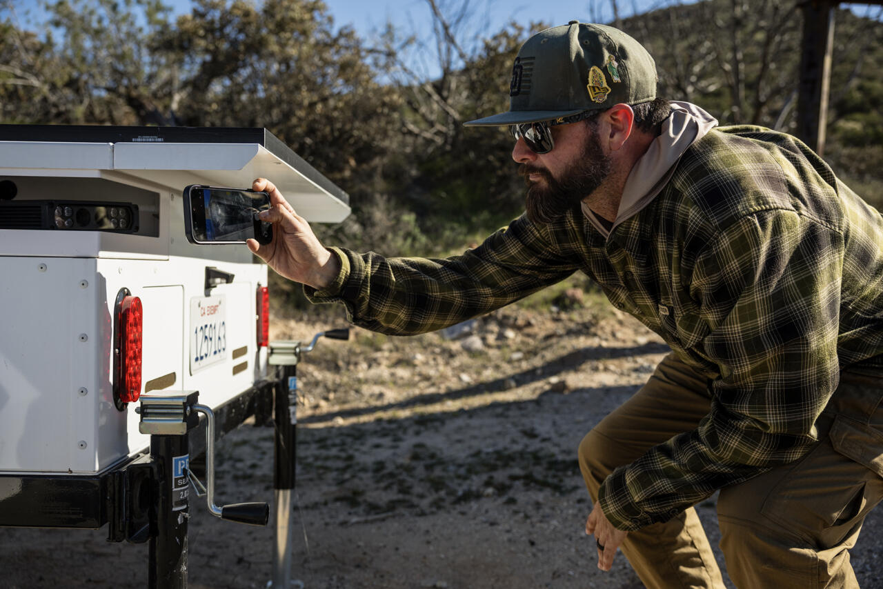 A person wearing a cap, sunglasses, and a plaid shirt crouches beside a roadside trailer device, holding a phone up to photograph or inspect its rear panel, with brush and dirt terrain in the background.